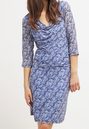 Frau in einem knielangen, blauen Kleid mit Blumenmuster, dreiviertellangen Ärmeln und drapiertem Ausschnitt vor einem einfarbigen Hintergrund.