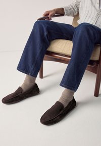 Chaussons brun foncé en daim avec une texture lisse et des détails cousus, portés avec des chaussettes côtelées beige et un jean en denim bleu.