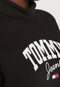 Czarna bluza z wysokim kołnierzem, która wyróżnia się dużą białą grafiką "TOMMY" z napisem "Jeans", miękka w dotyku, a na dole znajduje się mały kolorowy akcent logo.