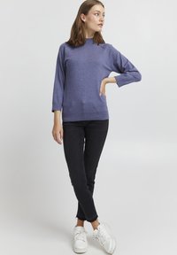 Zils džemperis ar augsto apkakli, trīs ceturtdaļas piedurknēm un ribotu malu; kombinēts ar melnām šaurām biksēm un baltām kedām.