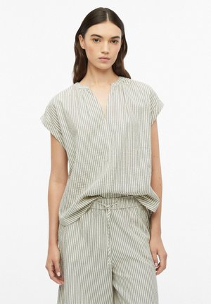 Vrouw draagt lichtgekleurde, verticaal gestreepte top met korte mouwen en bijpassende losse broek met koord in de taille, poserend tegen een witte achtergrond.