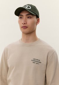 Joven con sudadera beige con el texto "LES DEUX COPENHAGEN EST. 2011" y gorra verde oscuro con un diseño bordado blanco.