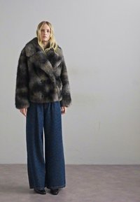 Płaszcz z sztucznego futra w odcieniach brązu i szarości z szerokim kołnierzem, zestawiony z luźnymi ciemnoniebieskimi spodniami w wzory i czarnymi, spiczastymi butami.