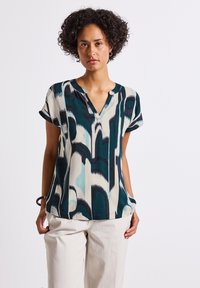 Model stands confidently, wearing a patterned blouse with teal, navy, and cream colors and light beige trousers, set against a plain background.