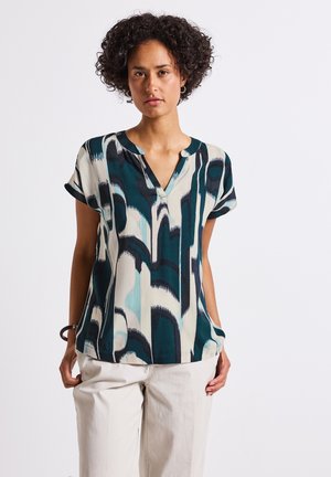 Das Model steht selbstbewusst da und trägt eine gemusterte Bluse in Türkis, Marineblau und Creme sowie eine hellbeige Hose, vor einem schlichten Hintergrund.