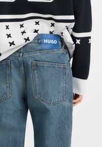 Jeans in denim azzurro chiaro con una tasca posteriore rettangolare, cuciture argento e un'etichetta blu con "HUGO" sulla vita.