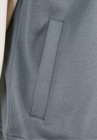 Gros plan sur une poche verticale à fente sur un vêtement en tissu gris, montrant les détails des coutures et la texture du matériau.