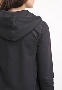Felpa nera con cappuccio realizzata in un tessuto morbido, dotata di cappuccio con cordino e schiena senza cuciture. Il design è semplice e casual, senza decorazioni visibili.