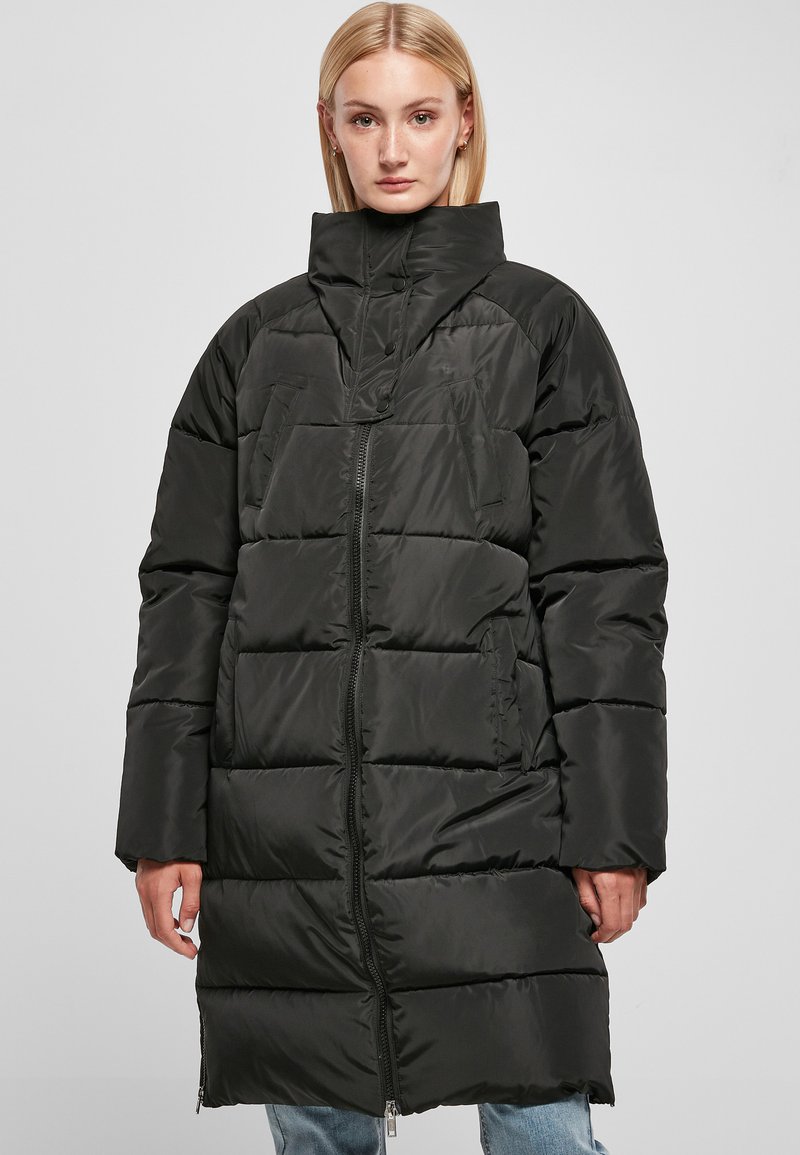 Cappotto trapuntato nero con colletto alto, caratterizzato da sezioni a righe orizzontali, tasche laterali e una cerniera a tutta lunghezza. Tessuto liscio e lucido.