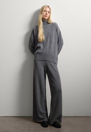 Maglione grigio a collo alto con vestibilità over, abbinato a pantaloni larghi grigi e scarpe slip-on nere, su uno sfondo neutro.