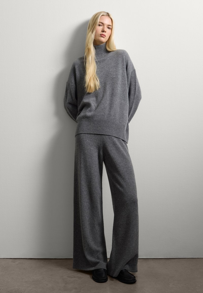 Maglione grigio a collo alto con vestibilità over, abbinato a pantaloni larghi grigi e scarpe slip-on nere, su uno sfondo neutro.