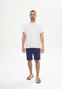 Weißes T-Shirt mit kurzen Ärmeln, kombiniert mit marineblauen Shorts und beigen Sandalen. Der Stoff wirkt glatt. Die Shorts haben einen elastischen Bund.