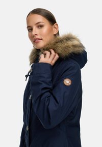 Navy blue parka with a faux fur-lined hood, featuring a button closure and a circular logo patch on the sleeve. Smooth, durable fabric.