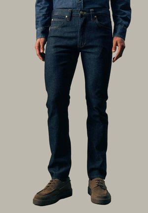 Mann trägt dunkelblaue Slim-Fit-Jeans, ein passendes Jeanshemd und braune Wildleder-Schnürschuhe, steht vor einem neutralen Hintergrund.
