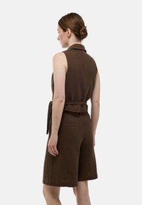 Jumpsuit sans manches côtelé marron avec un col, une ceinture nouée à la taille et des shorts, présentant un tissu texturé et des détails discrets à l'arrière.