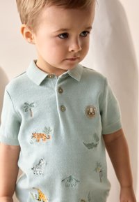 Hele sinine lühikeste varrukatega polo-särk tikanditega, millel on looma motiivid, sealhulgas lõvi ja palmipuud. Krae juures kaks pruuni nuppu.