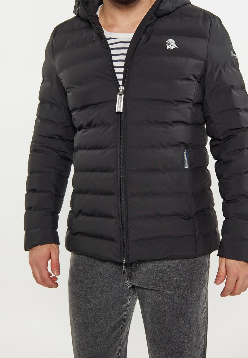 Homme portant une veste matelassée noire avec une capuche, unie à une chemise rayée et un jean gris, debout devant un fond blanc.