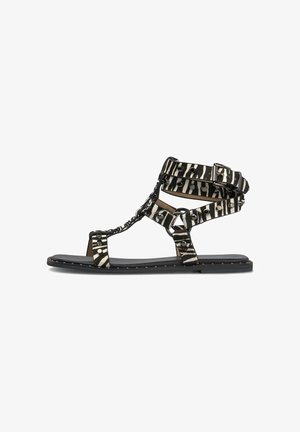 Sandale gladiateur plate avec des brides noir et blanc à rayures zébrées, ornées de clous métalliques et une fermeture à boucle à la cheville, sur une semelle noire.