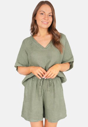 Woman with long brown hair wearing loose olive green short-sleeve top and matching shorts, standing with hands clasped at waist.