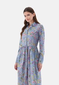 Abito floreale blu chiaro con maniche lunghe, chiusura a bottoni sul davanti e dettagli di gonna arricciata. Presenta motivi floreali colorati e cuciture visibili.