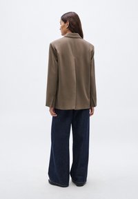 Καφέ σακάκι με προσαρμοσμένη ραφή και γιακά με μικρό άνοιγμα, με μία εγκοπή στην πλάτη, σε συνδυασμό με παντελόνι από σκούρο τζιν με φαρδιά μπατζάκια. Λείο υλικό.