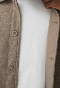 Braune Jacke aus Wollmischung mit glatter Textur, großen Knöpfen und hellfarbigem Innenfutter über einem weißen Hemd.