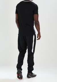 Czarna sportowa koszulka z białymi akcentami na ramionach, w połączeniu z czarnymi dresami z białymi paskami po bokach. Model ma na sobie czarne korki piłkarskie.