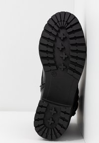 Semelle de botte noire à crampons profonds, poignées en forme de croix et indication de taille 37, appuyée contre un angle de mur et sol blancs.