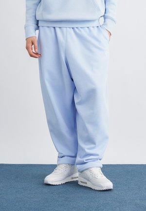 Persona vestida con pantalones deportivos azul claro y sudadera a juego, de pie sobre una alfombra azul, con zapatillas blancas y manos en los bolsillos.