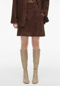 Jupe mini en daim marron avec une taille ceinturée, des poches latérales et une texture lisse, associée à des bottes beiges haute genou.
