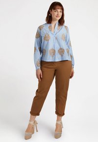 Hellblaue Bluse mit goldenen Löwenmotiven, kragenloses Design, lange Ärmel, kombiniert mit braunen hochgekrempelten Hosen und beigen Keilsandalen.