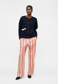 Sieviete stāv ar rokām kabatās, valkājot tumši zilu pogātu kardigānu un oranži baltas vertikālas svītras plašās biksēs.