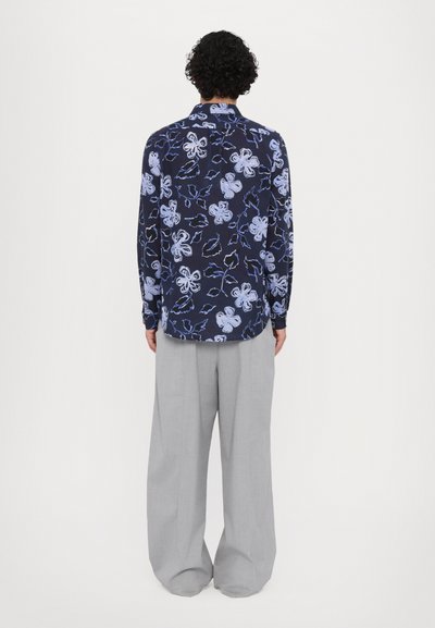 Camisa navy con un patrón floral en blanco y azul claro, de mangas largas y con un dobladillo curvado, combinada con pantalones de pierna ancha en gris claro.