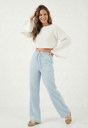 Uśmiechnięta kobieta z długimi włosami, ubrana w biały, długi rękaw krótki top, jasnoniebieskie szerokie spodnie i beżowe sandały, pozująca na jednolitym tle.