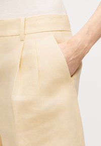 Χέρι εισαγόμενο στην εμπρόσθια τσέπη παντελονιού μεσαίου ύψους σε μπεζ χρώμα, με θηλιές ζώνης, φορεμένου με ανοιχτόχρωμο τοπ.