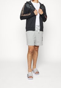 Svart zip-up hoodie med orange accentstrimmor, vit t-shirt, grå shorts och gråa slide-sandaler står på en enkel bakgrund.