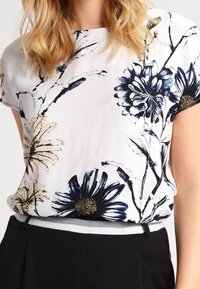 Blusa floreale bianca con accenti blu e oro, maniche corte, con disegni di fiori dipinti a mano e una vestibilità ampia. Tessuto di texture liscia.