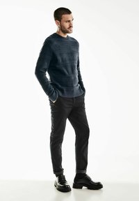 Pull en maille navy avec des rayures texturées, associé à un pantalon sombre et des chaussures noires massives, créant un look superposé.