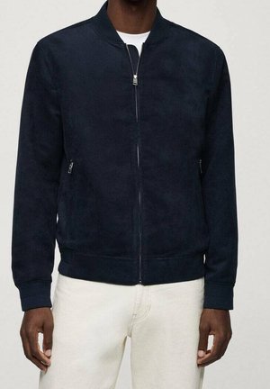 Man wearing a navy blue zip-up bomber jacket over a white shirt and light beige pants, standing against a plain background.