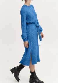 Robe midi bleue en tricot avec des manches longues, texture côtelée, ceinture à la taille et fentes latérales, associée  à des bottes noires à talons épais.
