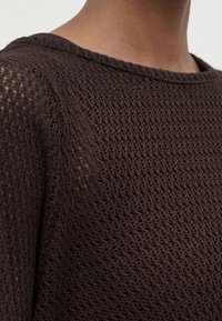 Gros plan d’un pull marron tricoté avec un motif en maille ajourée sur la partie supérieure de la poitrine et l’épaule d’une personne.