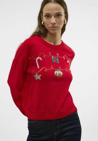 Maglione rosso lavorato a maglia con ricami festivi raffiguranti una canna di zucchero, regali e ornamenti su un filo dorato. Spalle scese e fondo a coste.