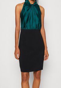 Satinová tyrkysová halenka s halter krkem a plisováním, doplněná o přiléhavou černou sukni, vytvářející elegantní siluetu. Hladký materiál, který nabízí kontrast barev.