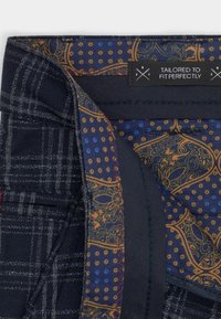 Elegantní kalhoty s námořnickým kostkovaným vnějším materiálem a propracovaným vzorovaným podšívkou v modré, zlaté a tmavých odstínech. Štítek hlásá "Ušito na míru."