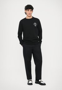 Czarna bluza crewneck z białą naszywką w kształcie rombu, zestawiona z czarnymi spodniami i czarnymi butami z białymi akcentami.