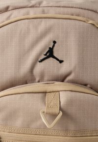 Mochila bege feita de tecido texturizado com o logótipo preto da Jordan, detalhes dourados e uma alça superior. Compartimentos com fecho visíveis.