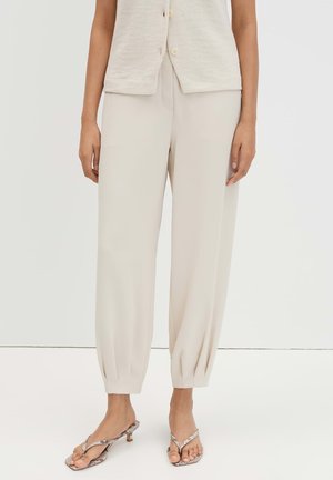 Unterkörper einer Person, die hellbeige, maßgeschneiderte Hosen mit aufgeschlagenen Säumen und offene, hochhackige Sandalen mit Schlangenledermuster trägt.