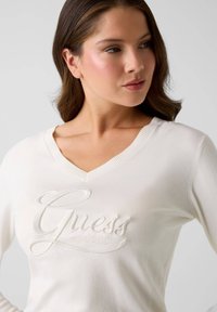 Weißer V-Ausschnitt-Pullover mit geripptem Saum und gesticktem "Guess est. 1981"-Logo. Weiche Textur; tailliertes Design betont Ausschnitt und Schultern.