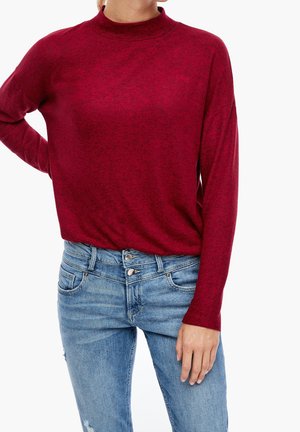 Kobieta ubrana w dopasowany bordowy sweter z długim rękawem i golfem, włożony do jasnoniebieskich dżinsów z podwójnym guzikiem.