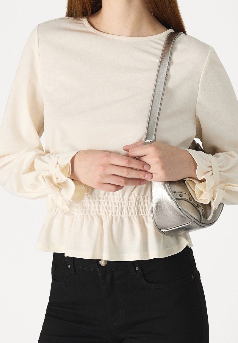 Femme portant un haut crème à manches longues avec une taille et des manches froncées, tenant un petit sac à bandoulière argenté métallique.